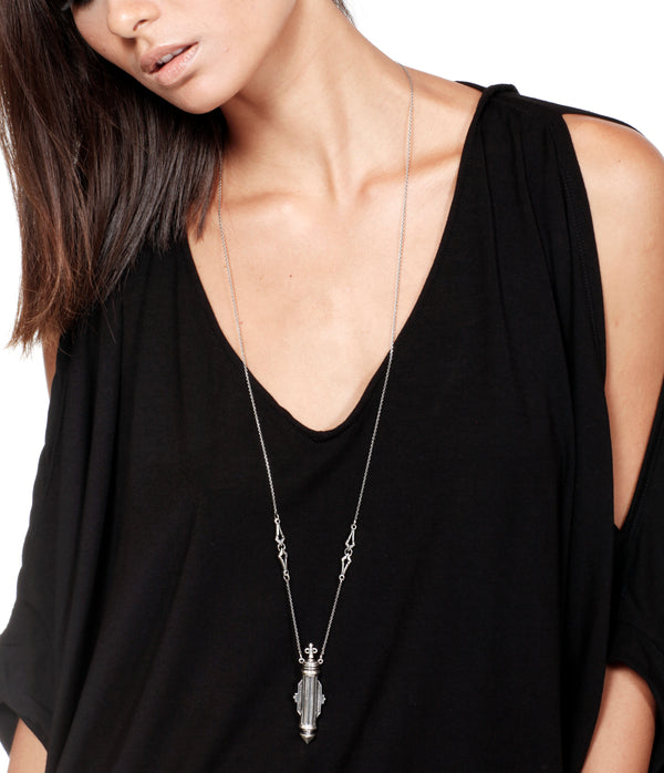 Person wearing a black top with a long silver necklace featuring a art nouveau perfume bottle pendant.