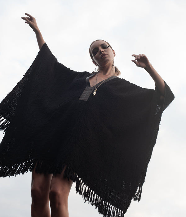 A person wearing a black, hand woven cotton poncho with a deep V-neck and leather trim, standing with arms raised and slightly bent.